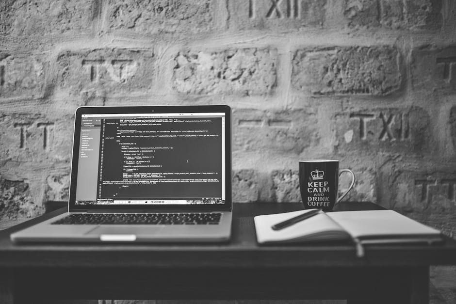 A black and white photograph of a workspace set up on a dark wooden desk, featuring an open laptop displaying lines of code on its screen, positioned centrally. To the right of the laptop, there is a