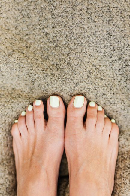 A close-up view of a pair of feet with well-maintained, neatly pedicured toenails, standing on a textured beige carpet in a well-lit room. The carpet surface appears clean and free of dust or stains.